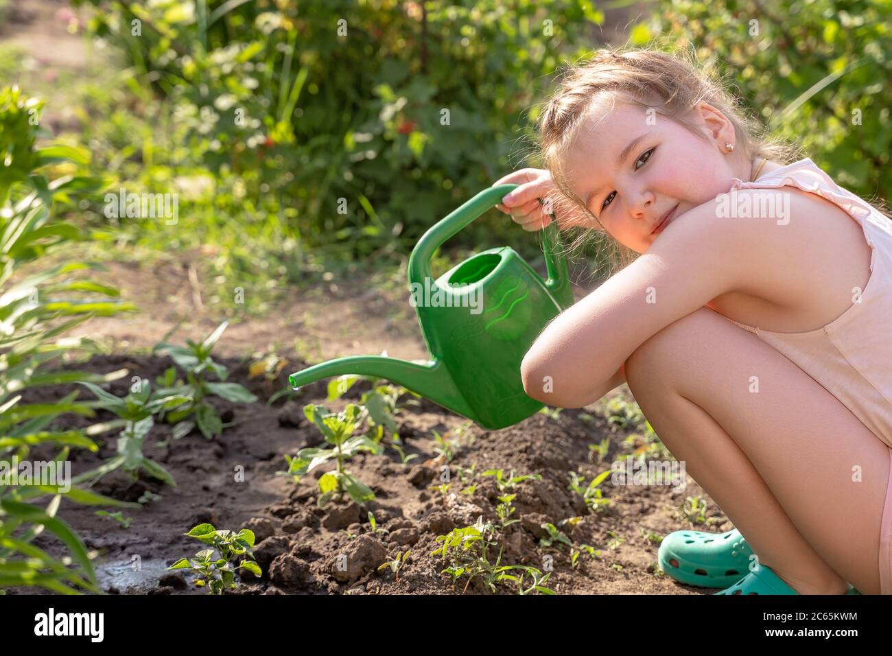Bella ragazza sorridente all'aperto in giardino si prende cura delle piante annaffiandola da annaffiatura verde lattina. Attività con bambini all'aperto. Foto Stock