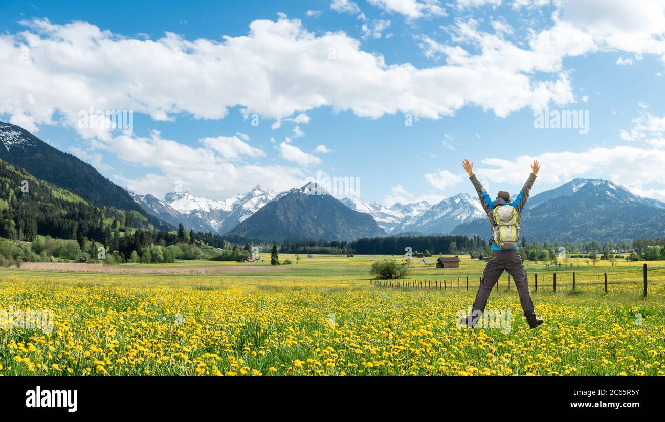 Hiker felice riuscito con il salto dello zaino con le braccia sollevate sul prato del fiore del dente di leone. Montagne innevate e casa tradizionale. Baviera, Alpi Foto Stock