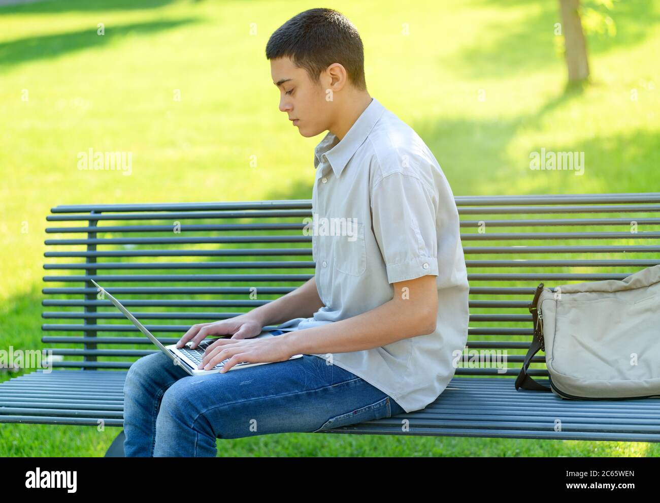 Ragazzo adolescente che usa il suo laptop nel parco mentre si siede su una panchina battendo con esso bilanciato sul suo grembo in una giornata estiva soleggiato contro un erba verde Foto Stock