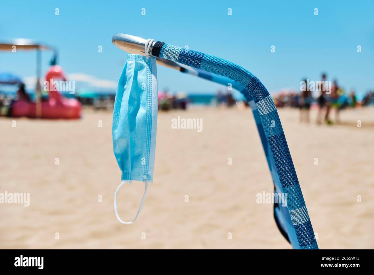 primo piano di una maschera chirurgica appesa su una sedia blu sulla spiaggia, con il mare sullo sfondo Foto Stock
