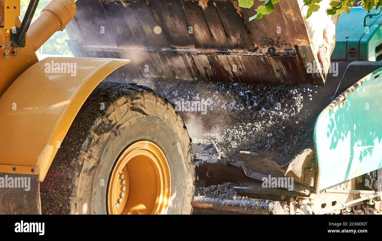 Il trattore a benna versa l'asfalto caldo in una livellatrice, costruendo una nuova strada in una città, materiale per la costruzione di strade da vicino. Foto Stock