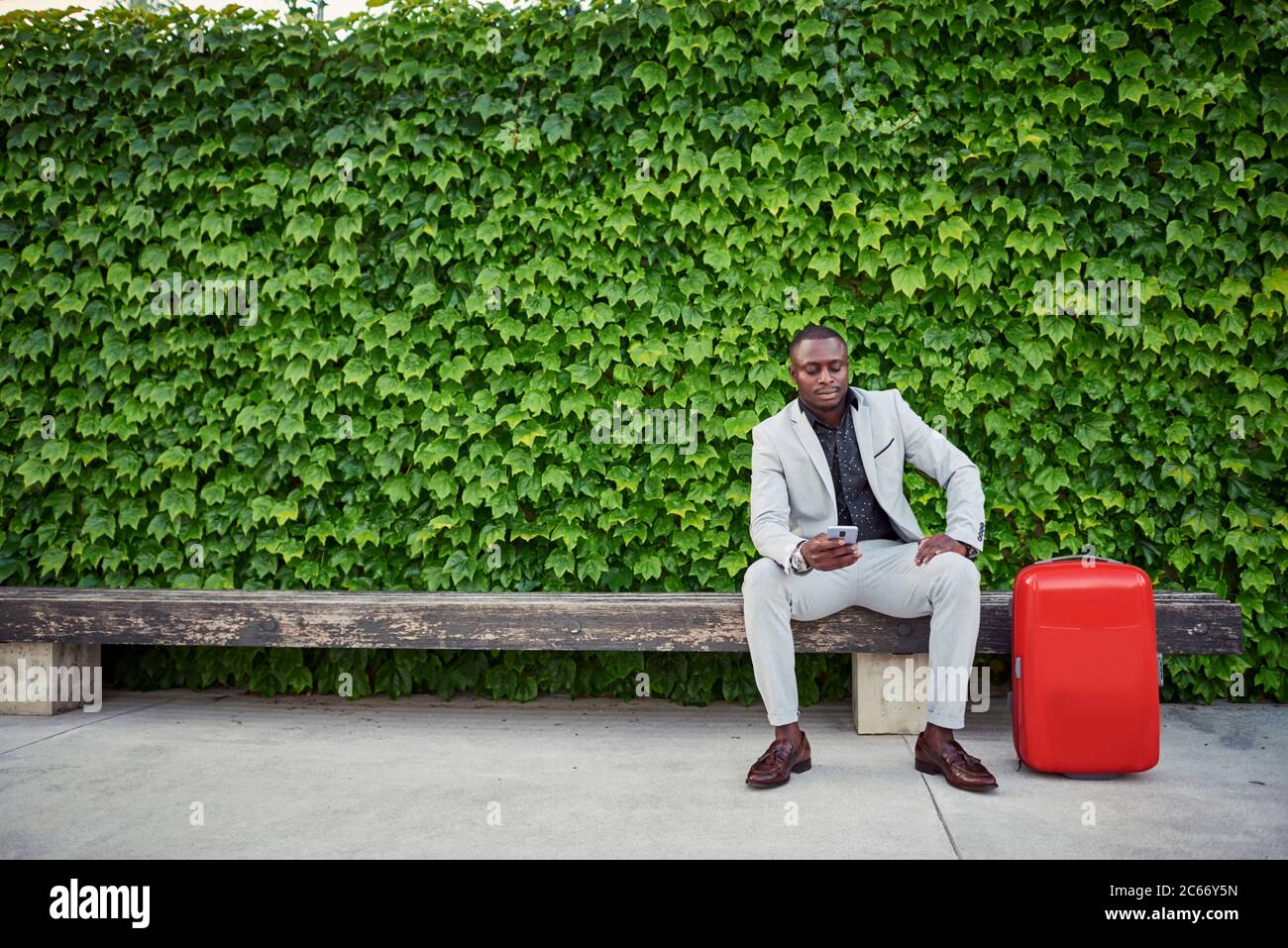 uomo d'affari seduto su una panchina con una valigia rossa e un telefono cellulare in mano. uomo d'affari in viaggio. Foto Stock