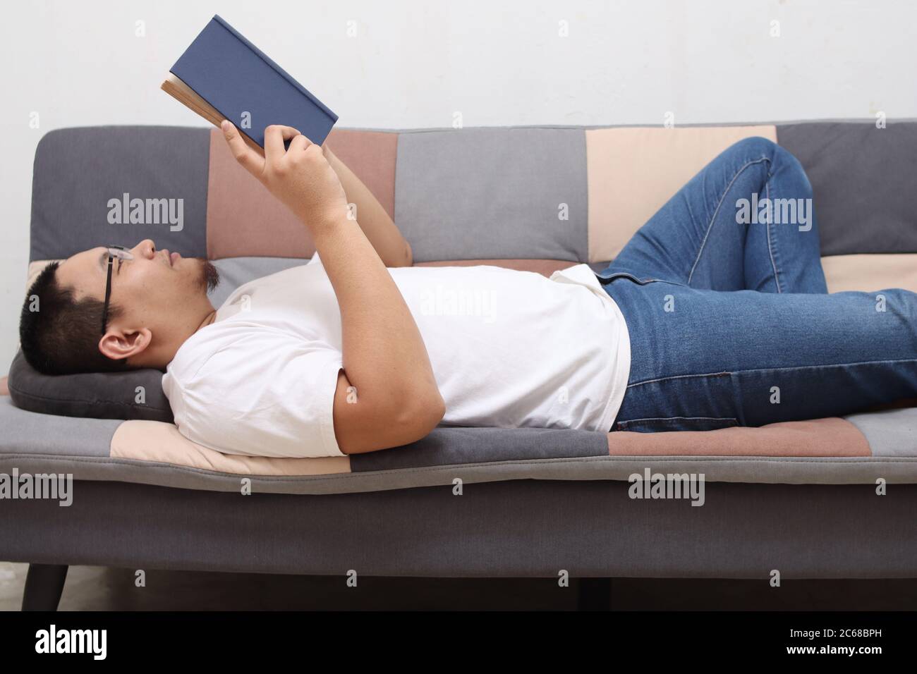 Felice uomo asiatico leggere un libro mentre si rilassa sul divano, godere week-end con attività di svago Foto Stock