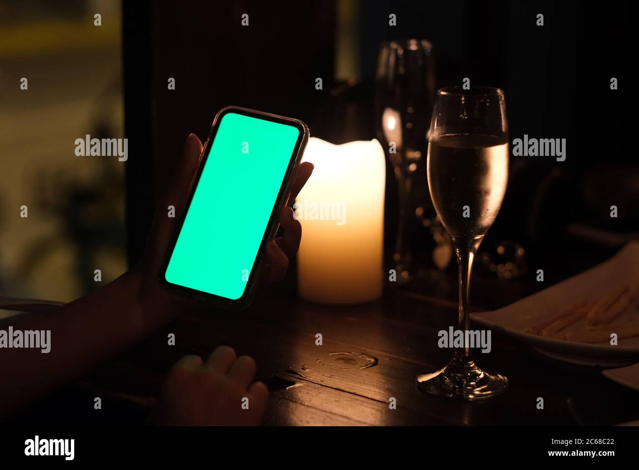 primo piano con una mano che mostra lo smartphone con schermo verde al bar di notte. bicchiere di vino e candela sfocati come sfondo Foto Stock