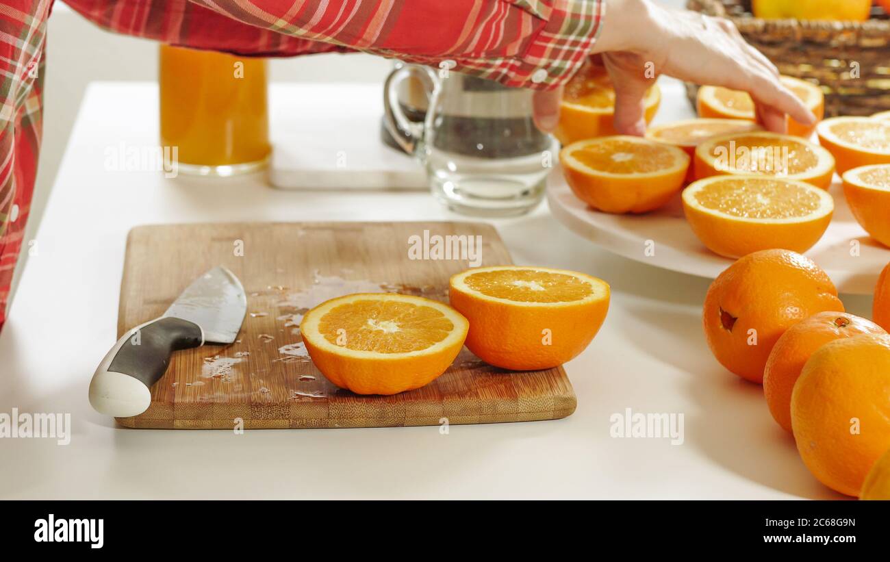 Donna taglia arance su una tavola da cucina in legno con sfondo bianco. Preparazione di succo d'arancia fresco con una centrifuga elettrica. Foto Stock
