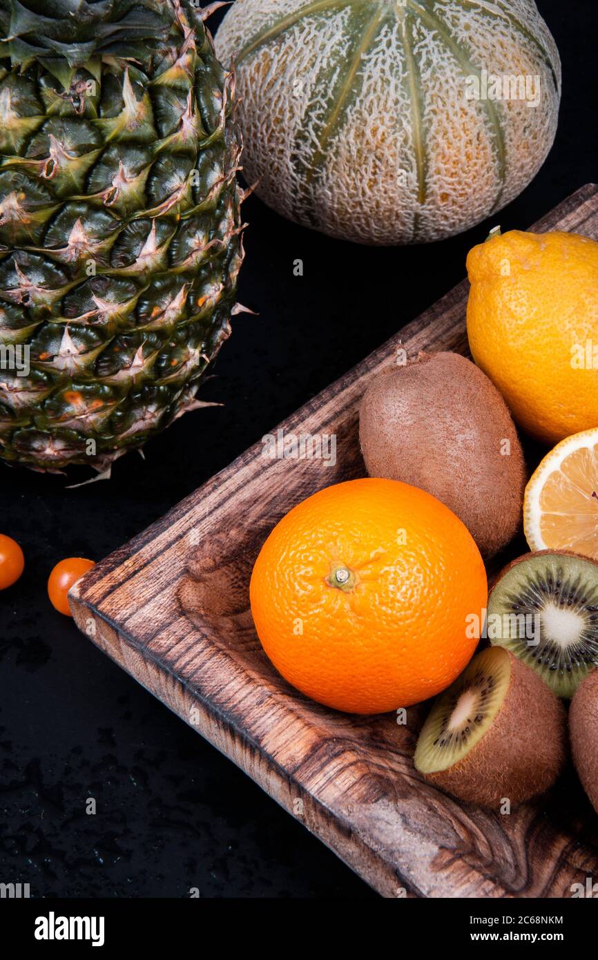 Una ciotola di legno piena di kiwi verdi, arance e limoni vicino ad un melone di miele, una mela di pino e una lanterna cinese su uno sfondo nero. Foto Stock