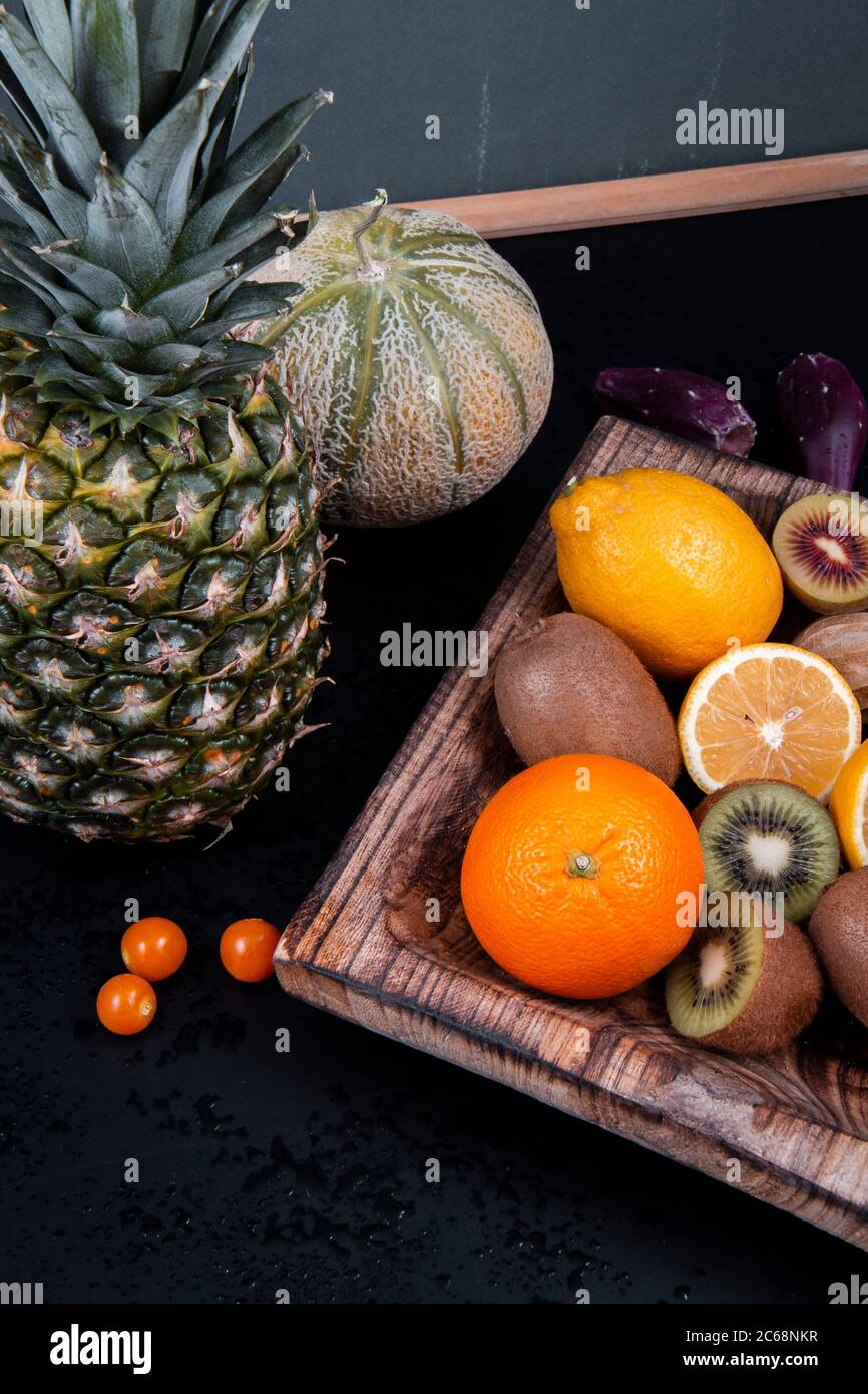 Una ciotola di legno piena di kiwi verdi, arance e limoni vicino ad un melone di miele, una mela di pino e una lanterna cinese su uno sfondo nero. Foto Stock