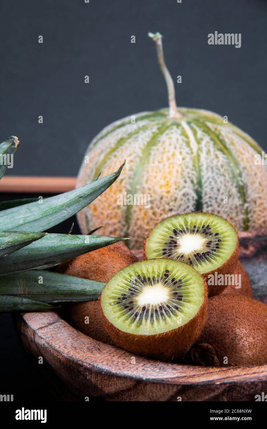 Una ciotola di kiwi pieno e dimezzato e una melone di melone di miele vicino alle foglie di un ananas. Foto Stock