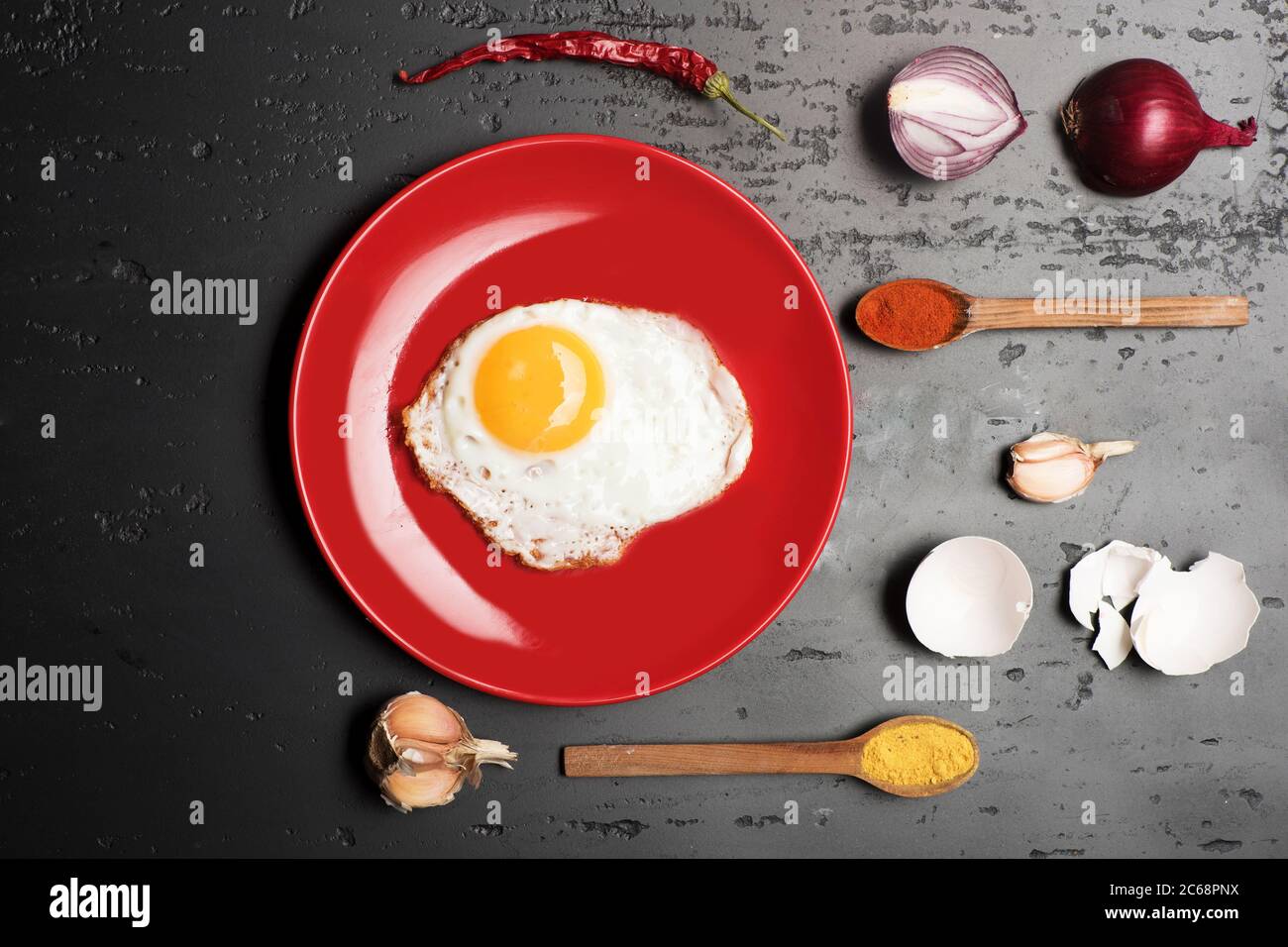 Cibo posto su piatto rosso su sfondo scuro. Omelette messa al centro del piatto. Condire con l'uovo fritto vicino a cucchiai con spezie, aglio, cipolla, gusci di uova e peperoncino rosso. Colazione e concetto di arte alimentare Foto Stock