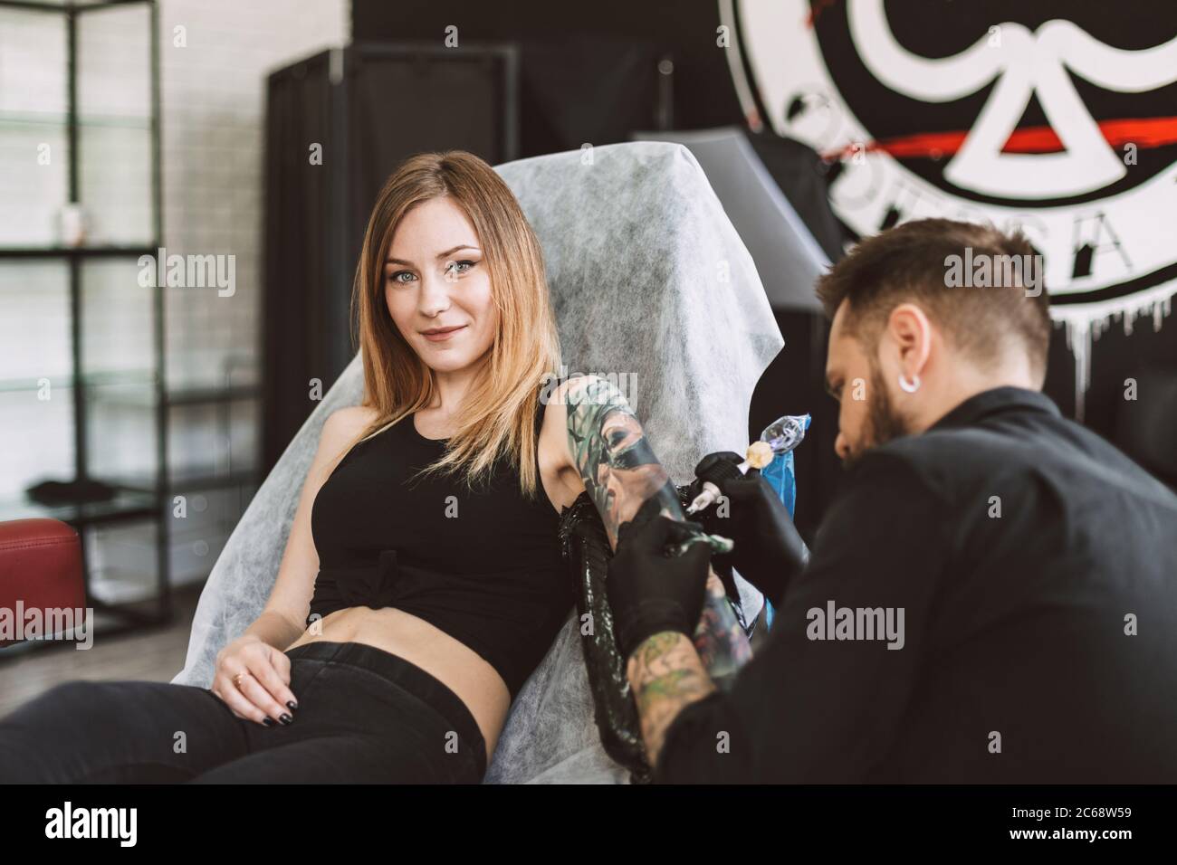 Sorridente tatuata ragazza che cerca in macchina fotografica professionale tatuaggio facendo il tatuaggio a mano utilizzando la macchina tatuaggio in studio Foto Stock