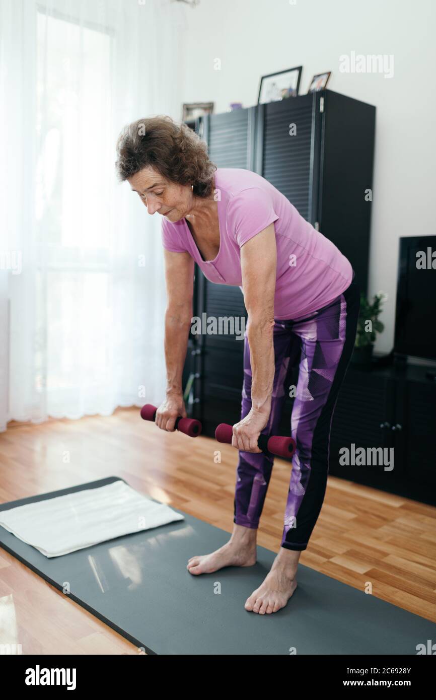 Donna anziana attiva che fa l'allenamento a casa. Foto a lunghezza intera di una donna di 70 anni focalizzata in viola sport abbigliamento allenamento la schiena con manubri bo Foto Stock