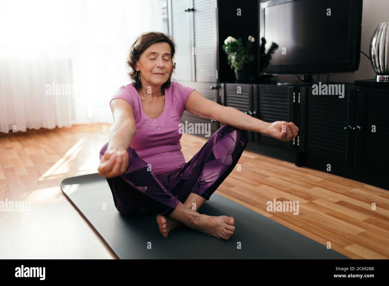 Donna senior focalizzata che fa yoga a casa. donna di 70 anni seduta su un tappetino fitness con mani distese che si riposano sulle ginocchia eseguendo yoga in liv Foto Stock