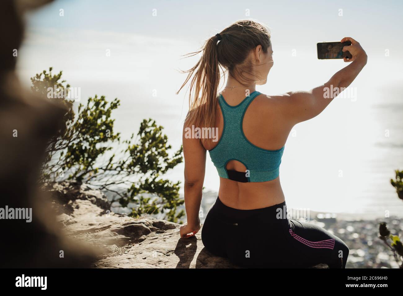 Vista posteriore di una giovane donna in forma seduta sulla scogliera che prende selfie. Donna in abbigliamento sportivo che fa un ritratto di auto con il telefono cellulare sulla cima della montagna. Foto Stock