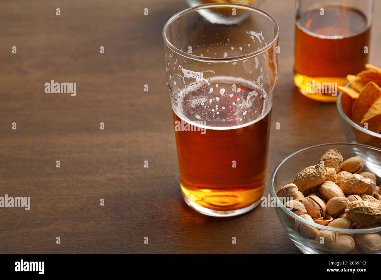 Bevanda del cliente. Bicchiere di birra e bottiglia incompiuto, pistacchio, patatine in piatti di vetro su tavolo di legno Foto Stock