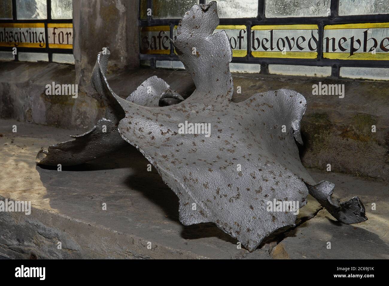 La cortina ritorta di shrapnel metallico, ora esposta sul davanzale nella Parrocchia di San Laurence a Hawkhurst, Kent, Inghilterra, è il resto della bomba volante tedesca V1 che è esplosa nel cimitero nell'agosto 1944, durante la seconda guerra mondiale, soffiando fuori molto antico vetro macchiato e causando ulteriori danni estensivi. Sebbene la chiesa non fu completamente riparata fino al 1957, pezzi di vetro frantumato furono successivamente fatti in un nuovo tondo e beneficiò anche di donazioni di vetro antico da altrove. Foto Stock