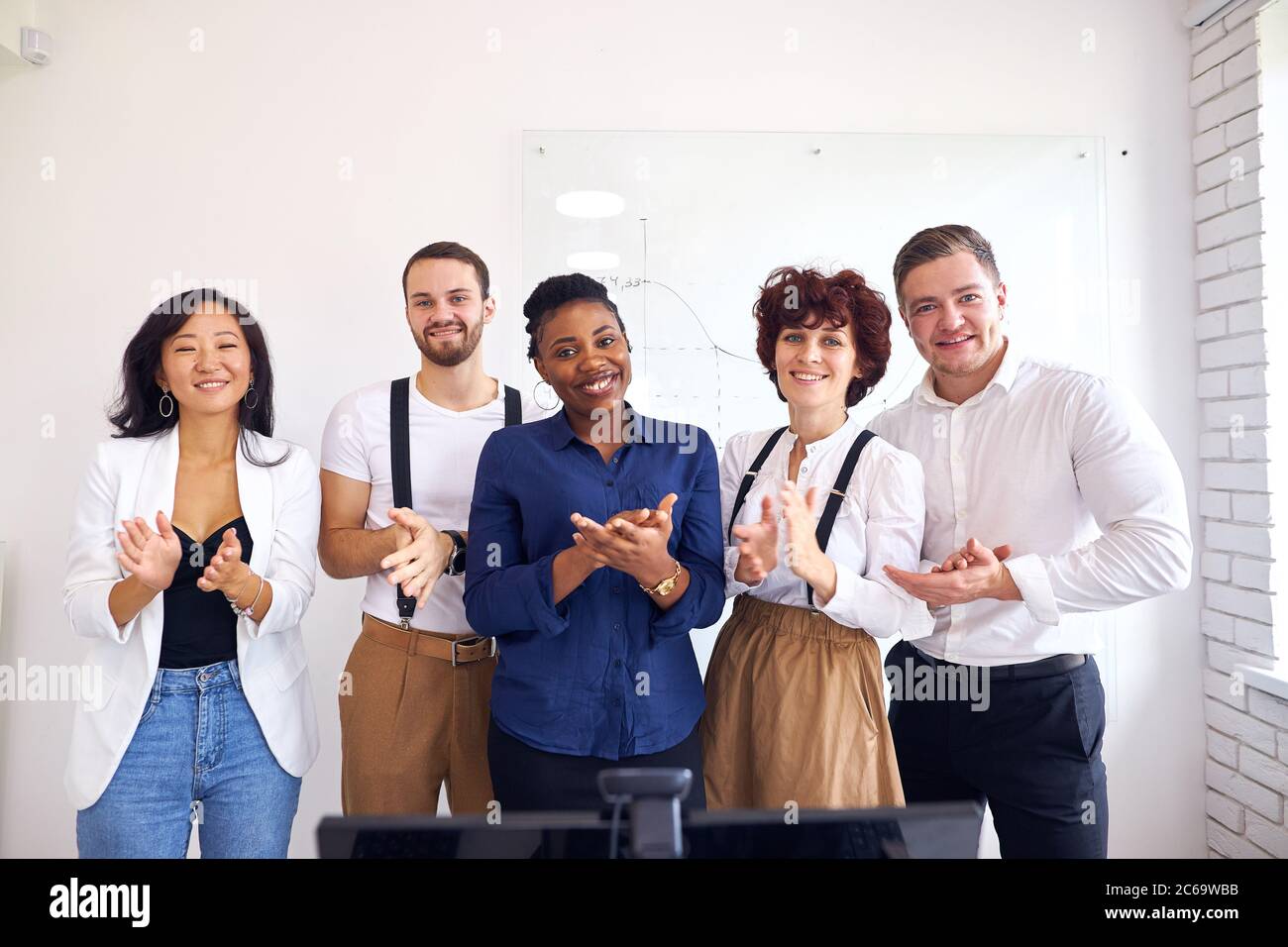 Sicuro sorridente gruppo multi-etnico loo alla macchina fotografica e applaudente, sfondo bianco lavagna a fogli mobili Foto Stock