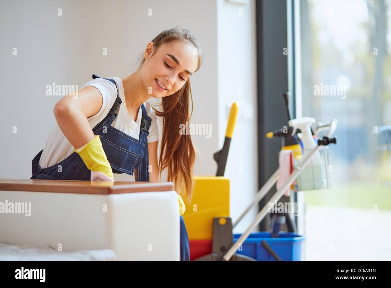 Ragazza caucasica positiva in attrezzatura speciale per pulire accuratamente la polvere da divano bianco in casa nuova. Fondo detergenti Foto Stock