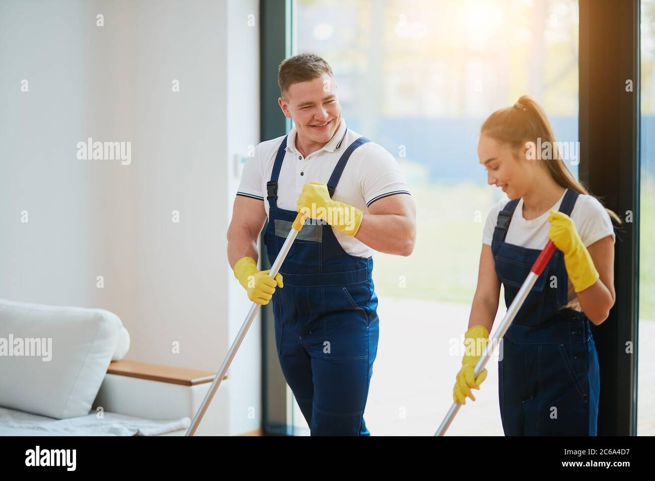 Il personale del servizio di pulizia lavora insieme, lavando il pavimento con la mop. Indossare indumenti speciali per la pulizia. Sfondo panoramico della finestra Foto Stock