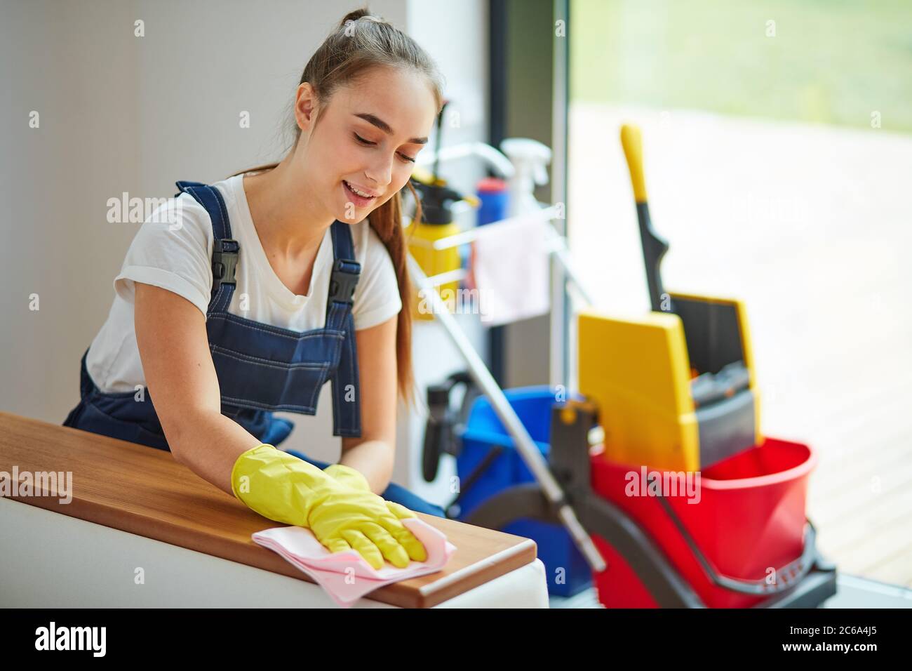 Ragazza caucasica positiva in attrezzatura speciale per pulire accuratamente la polvere da divano bianco in casa nuova. Fondo detergenti Foto Stock