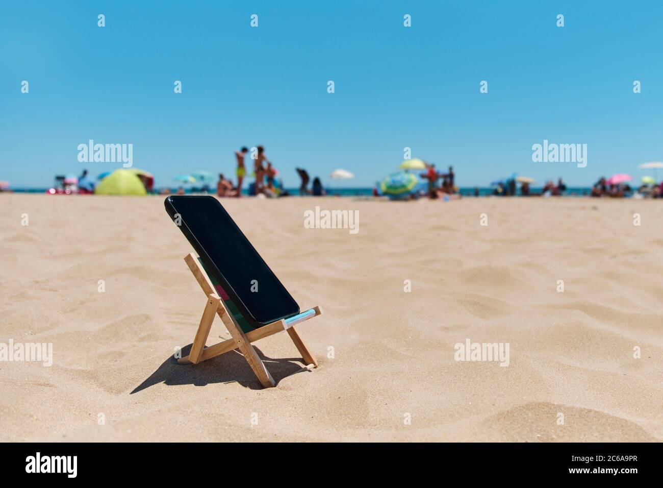 uno smartphone nero su una sedia a sdraio sulla sabbia della spiaggia, raffigurante il concetto di detox digitale, con persone irriconoscibili godendosi e il mare in t Foto Stock
