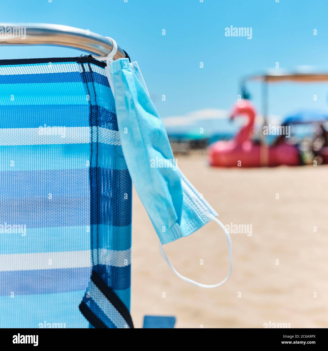 primo piano di una maschera chirurgica appesa su una sedia blu sulla spiaggia, con il mare sullo sfondo Foto Stock