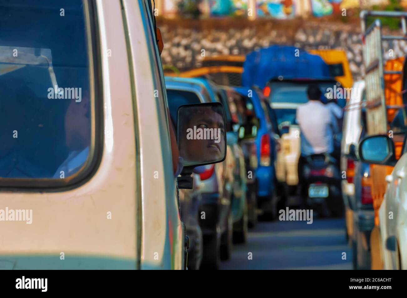 Il riflesso del volto del conducente visibile sullo specchietto retrovisore di un pulmino Maruti. Una lunga fila di veicoli può essere vista intrappolata in un ingorgo stradale. Foto Stock