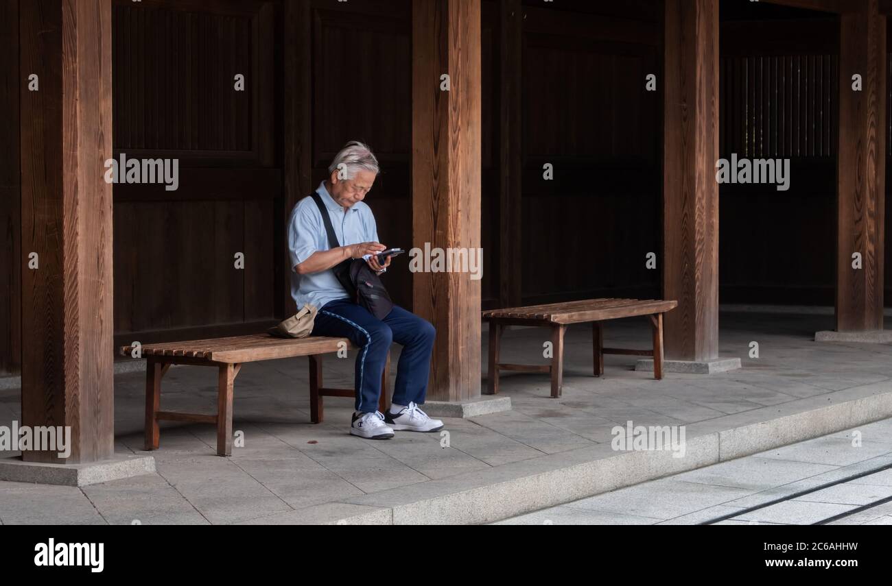 Anziano giapponese seduto su una panchina di legno con smartphone in Meiji Jingu interno composto, Tokyo, Giappone Foto Stock