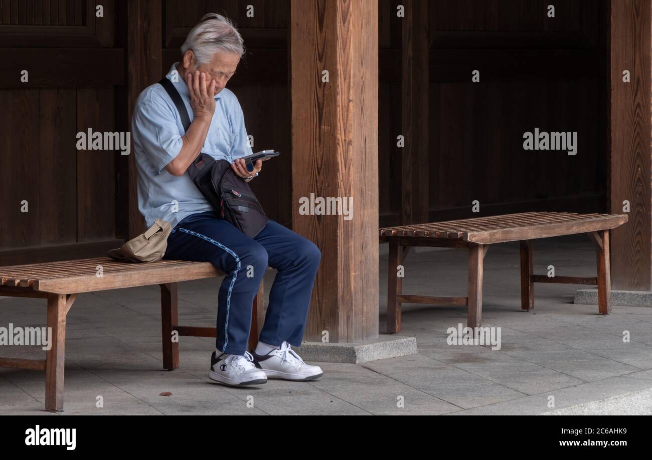 Anziano giapponese seduto su una panchina di legno con smartphone in Meiji Jingu interno composto, Tokyo, Giappone Foto Stock