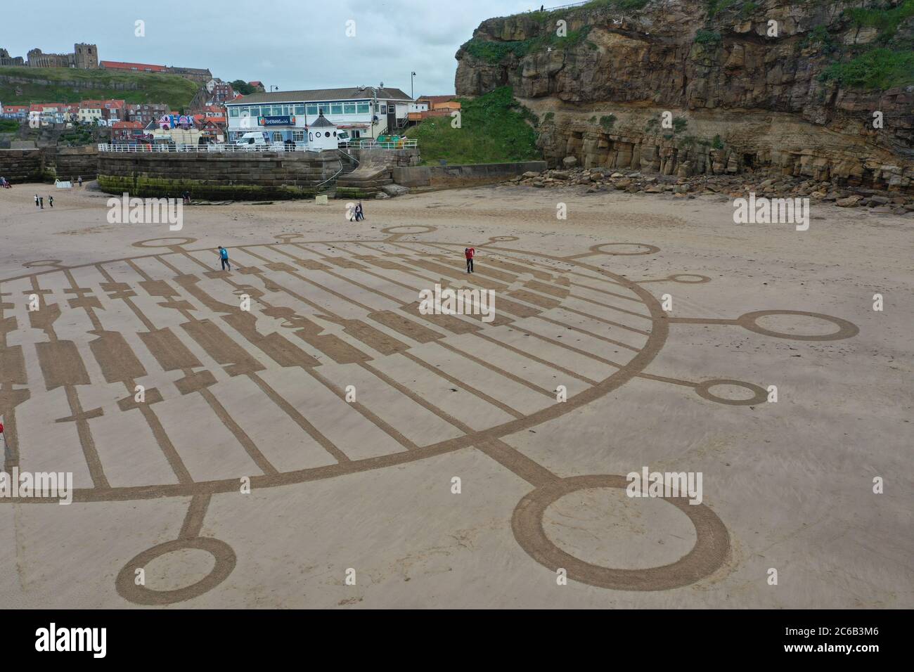 La gente cammina attraverso un pezzo gigante di arte della sabbia che è stato creato sulla spiaggia a Whitby, North Yorkshire, per aiutare a guidare a casa l'importanza di distanziamento sociale per coloro che visitano il mare e altri luoghi popolari. Foto Stock
