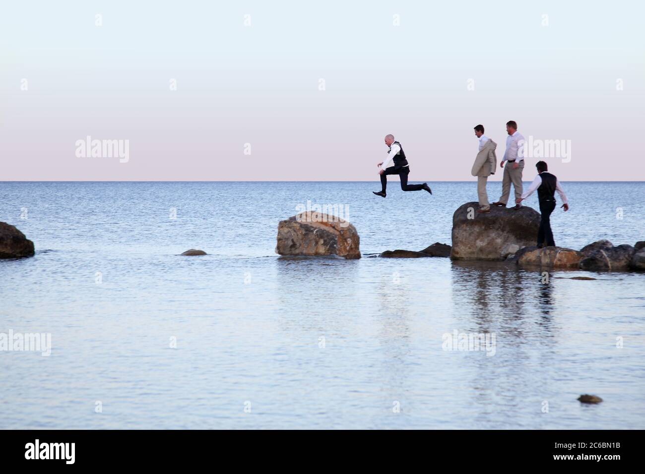 L'uomo compie un salto di fede tra le rocce in mare. Altri uomini in fila per saltare Foto Stock