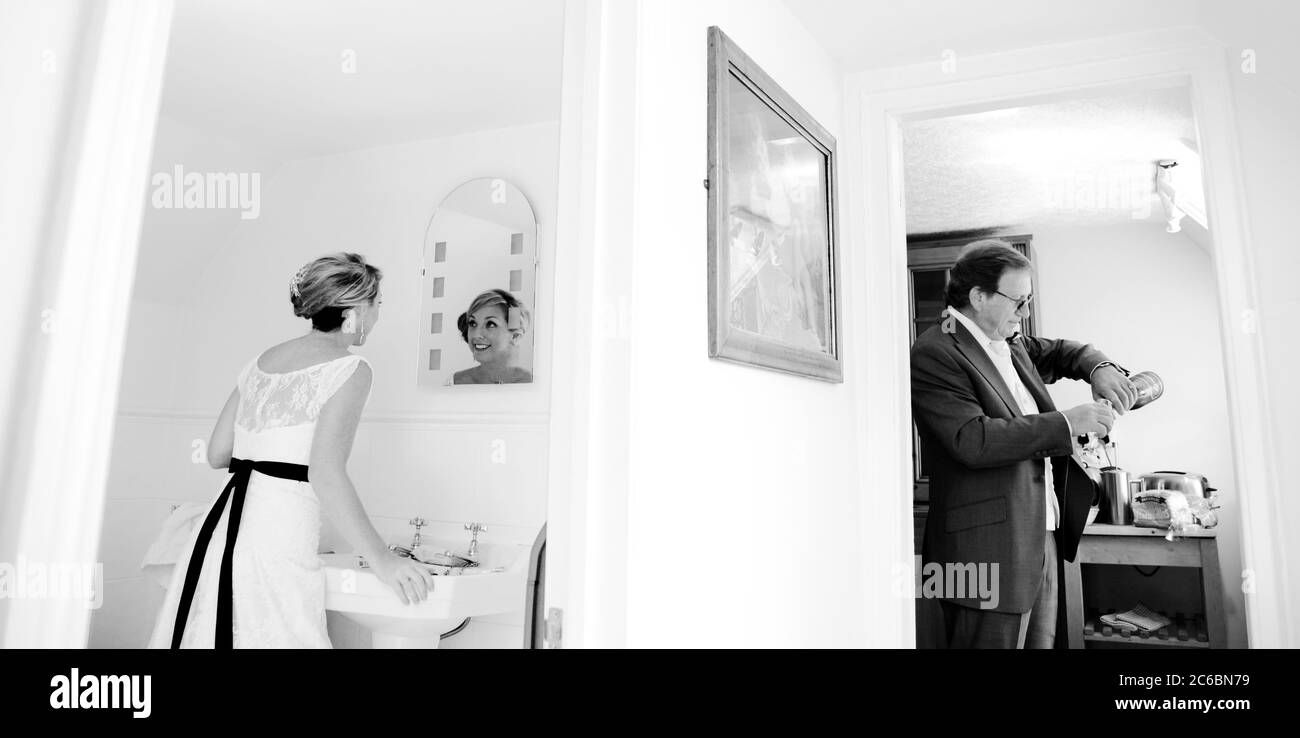 Sposa, anni trenta, si prepara davanti allo specchio da bagno mentre Padre della Sposa, anni cinquanta/sessanta, si versa una bevanda di champagne. Foto Stock