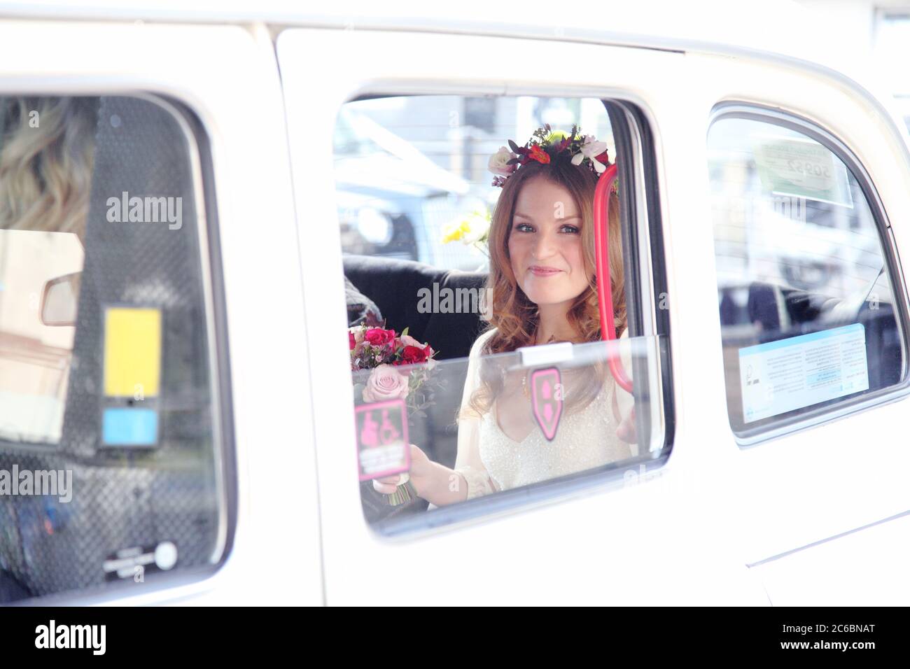 Anni venti rosso boho sposa in abito da sposa con ghirlande in rosso zenzero capelli tenendo un bouquet di fiori arriva al suo matrimonio in un taxi bianco. Foto Stock