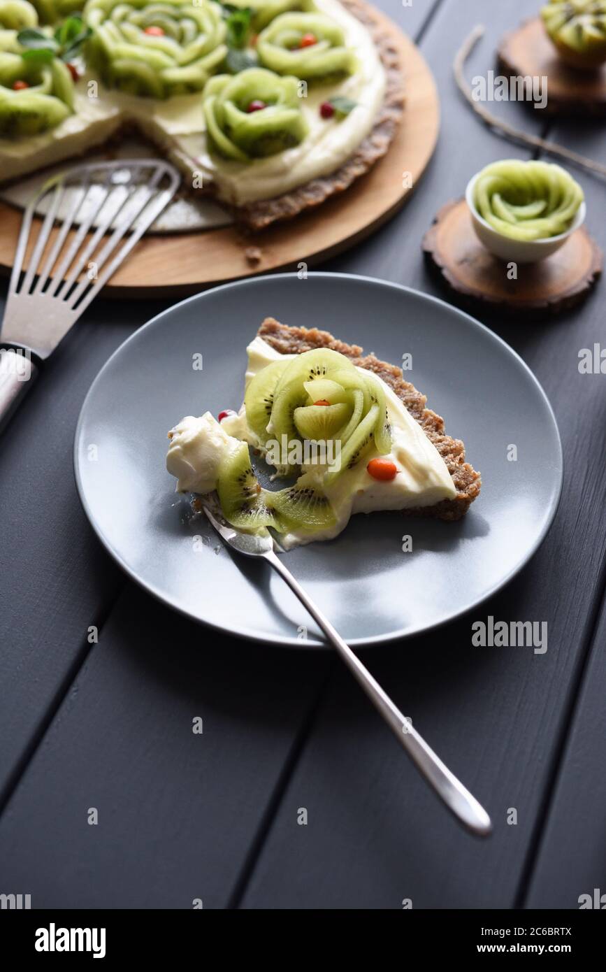 Dessert vegano crudo. Torta fatta in casa con rose kiwi, bacche e crema ripieno su crosta di noce su sfondo scuro con copyspace Foto Stock