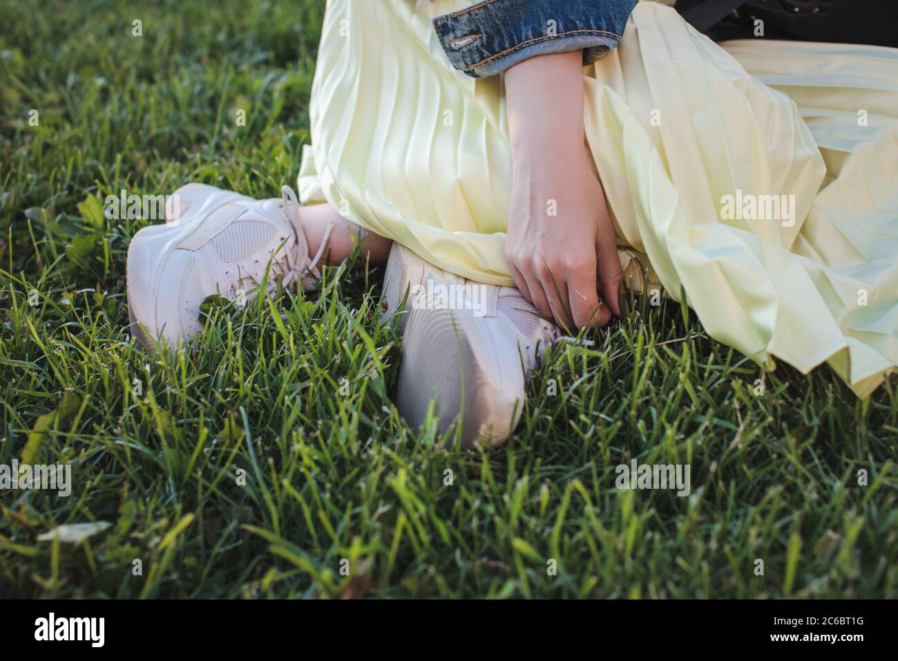 Una ragazza in sneakers sportivi bianchi e una gonna gialla si siede sull'erba nel parco. Cosce e mani closeup. Foto Stock