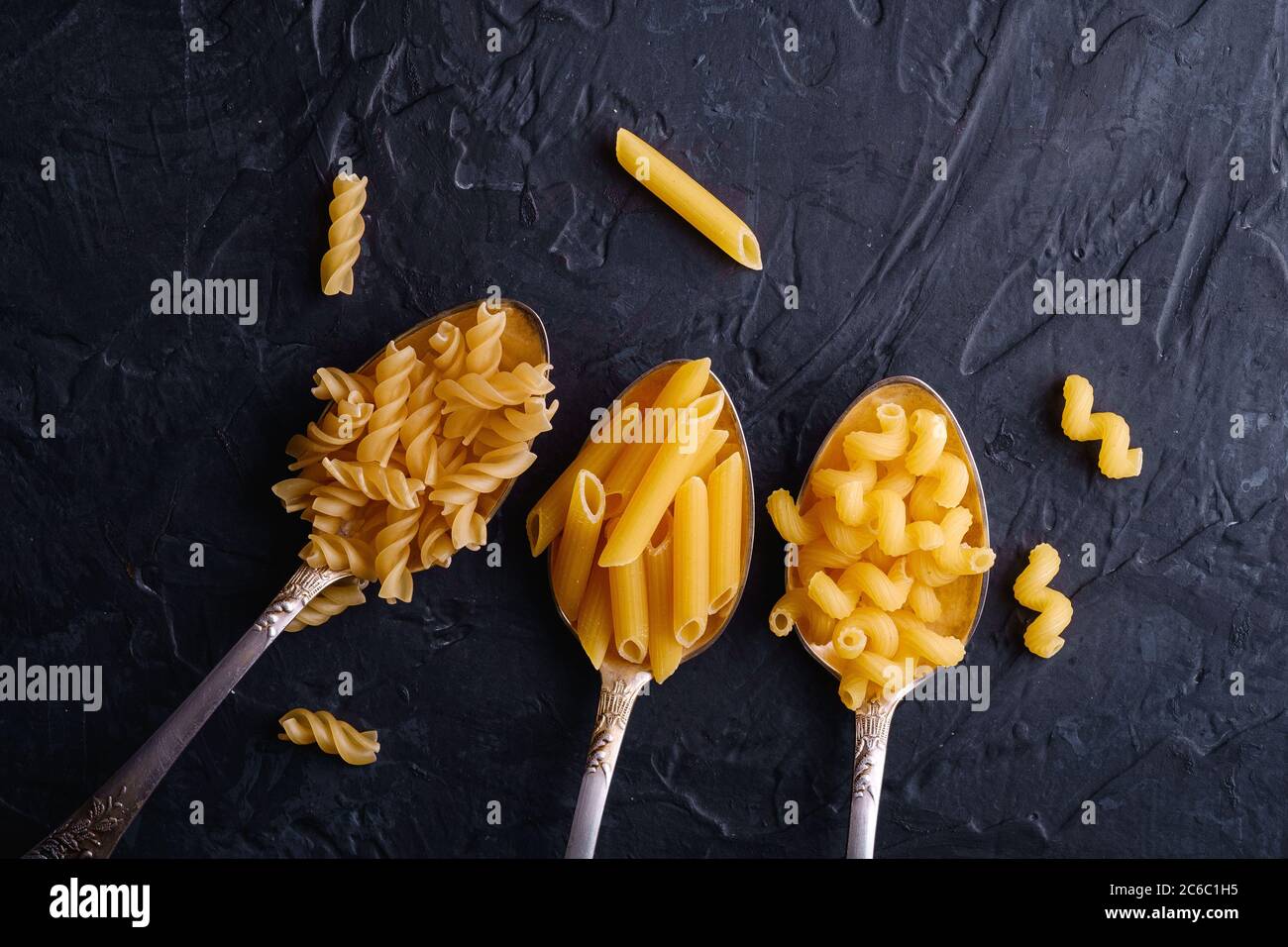 Tre cucchiai di posate con una varietà di pasta di grano dorato crudo su sfondo nero scuro testurizzato, vista dall'alto Foto Stock