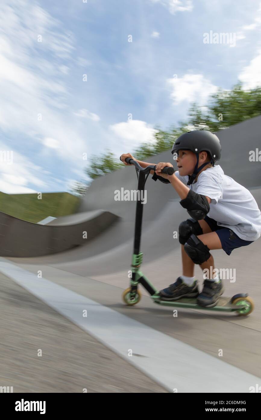 Un simpatico ragazzino guida uno scooter in uno skatepark. Un giovane atleta novizio trascorre del tempo libero in sport estremi. Foto di movimento, blu di movimento intercambiabile Foto Stock