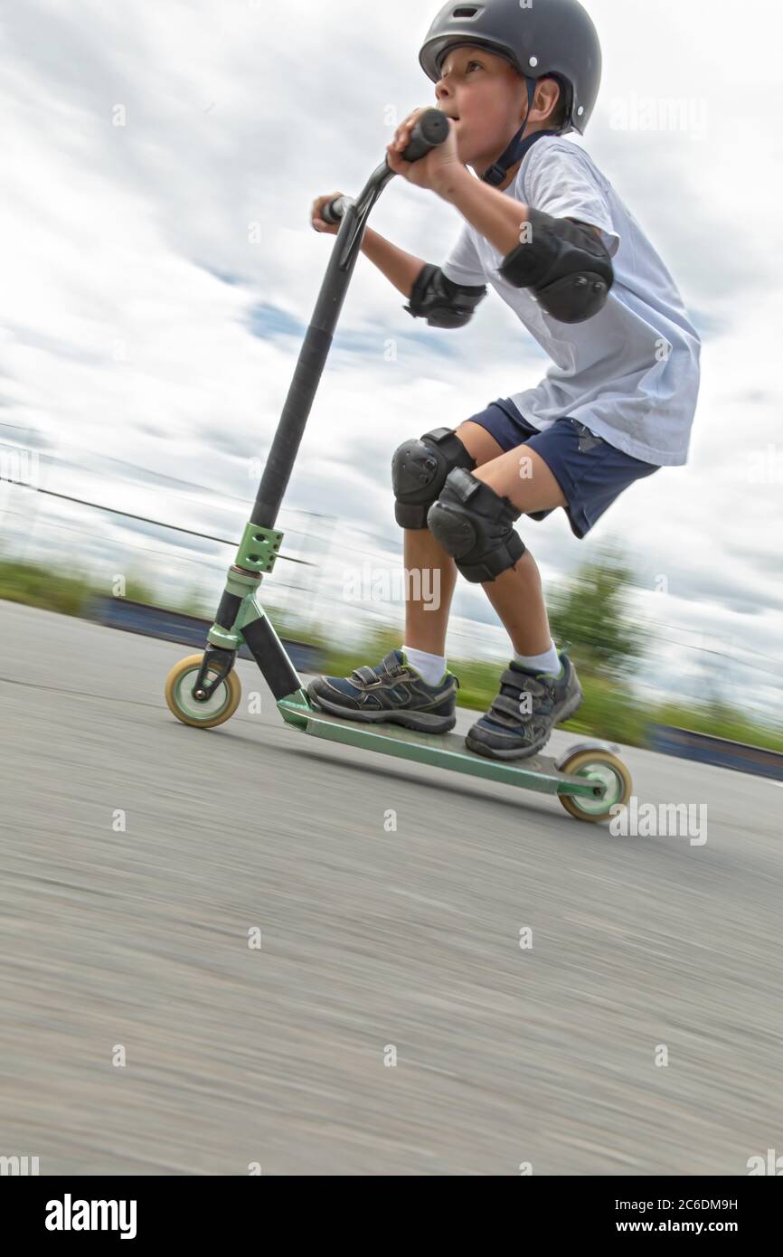 Un simpatico ragazzino guida uno scooter in uno skatepark. Un giovane atleta novizio trascorre del tempo libero in sport estremi. Foto di movimento, blu di movimento intercambiabile Foto Stock