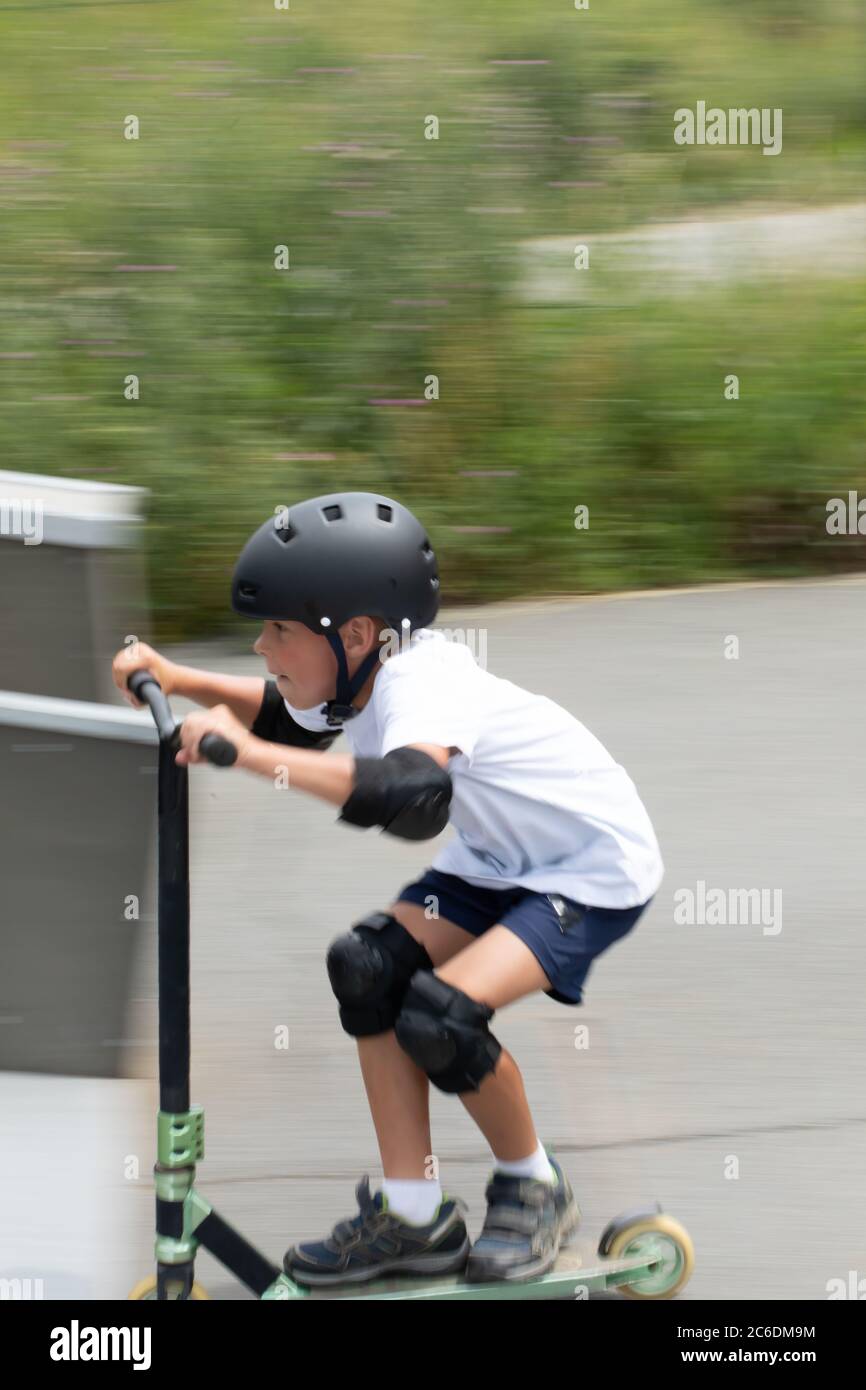 Un simpatico ragazzino guida uno scooter in uno skatepark. Un giovane atleta novizio trascorre del tempo libero in sport estremi. Foto di movimento, blu di movimento intercambiabile Foto Stock