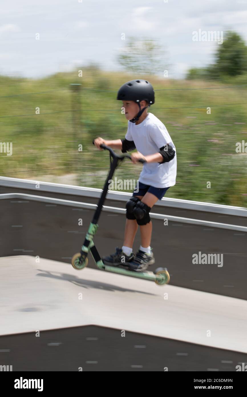 Un simpatico ragazzino guida uno scooter in uno skatepark. Un giovane atleta novizio trascorre del tempo libero in sport estremi. Foto di movimento, blu di movimento intercambiabile Foto Stock