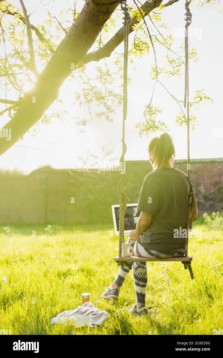 Donna con laptop che parla in swing in giardino soleggiato Foto Stock