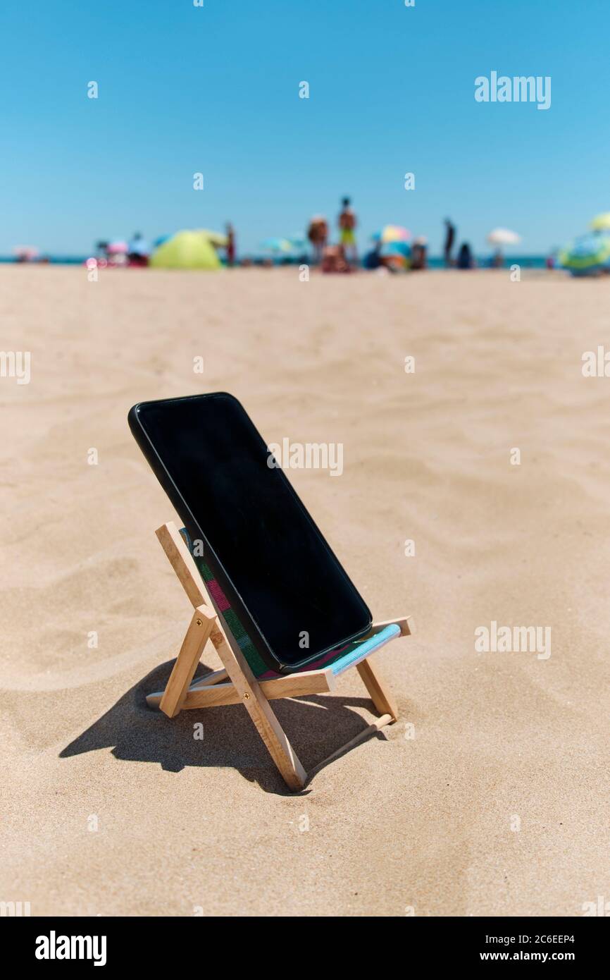 uno smartphone nero su una sedia a sdraio sulla sabbia della spiaggia, raffigurante il concetto di detox digitale, con persone irriconoscibili godendosi e il mare in t Foto Stock