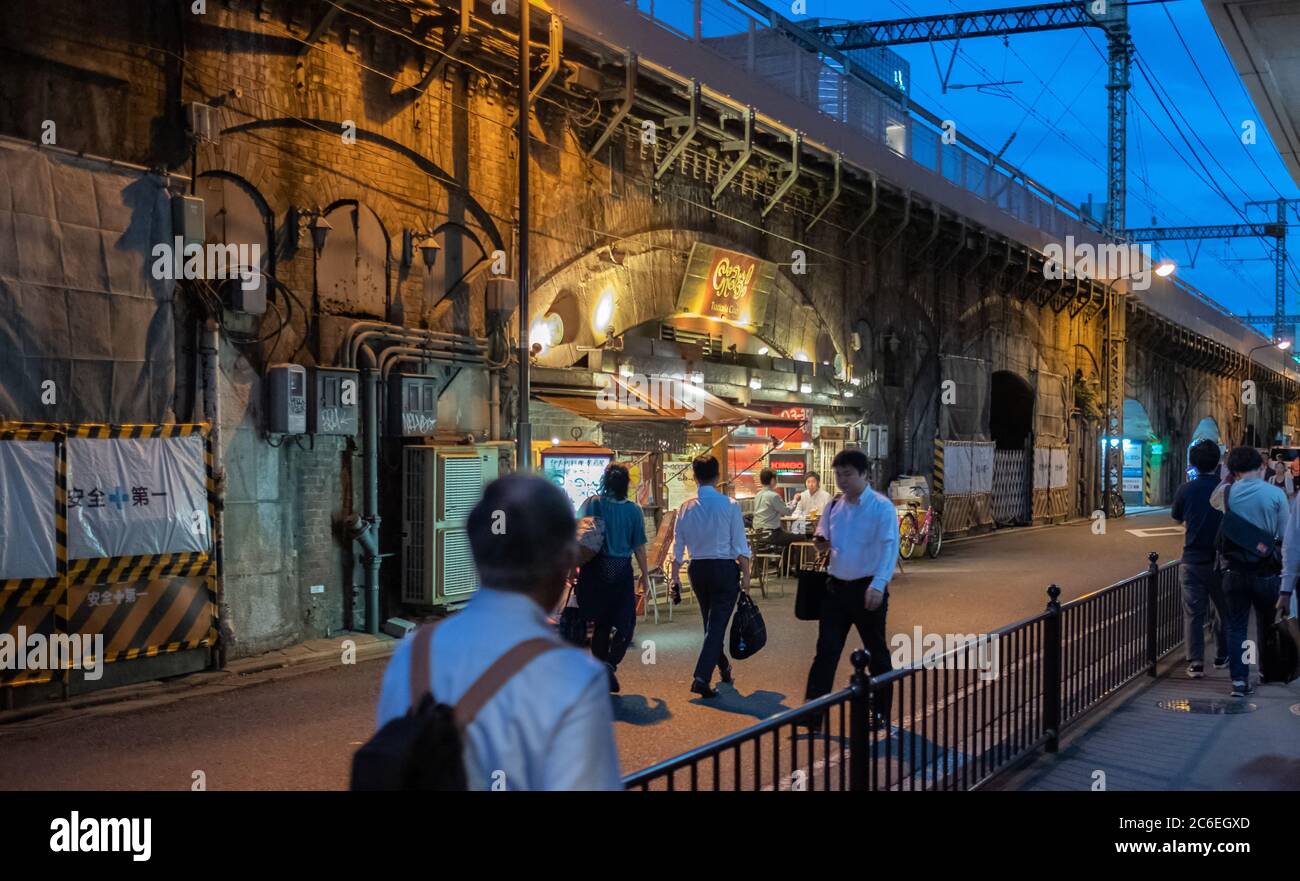 Pedoni che camminano lungo una stretta corsia posteriore accanto a una linea ferroviaria sopraelevata a Yurakucho, Tokyo, Giappone al tramonto. Foto Stock