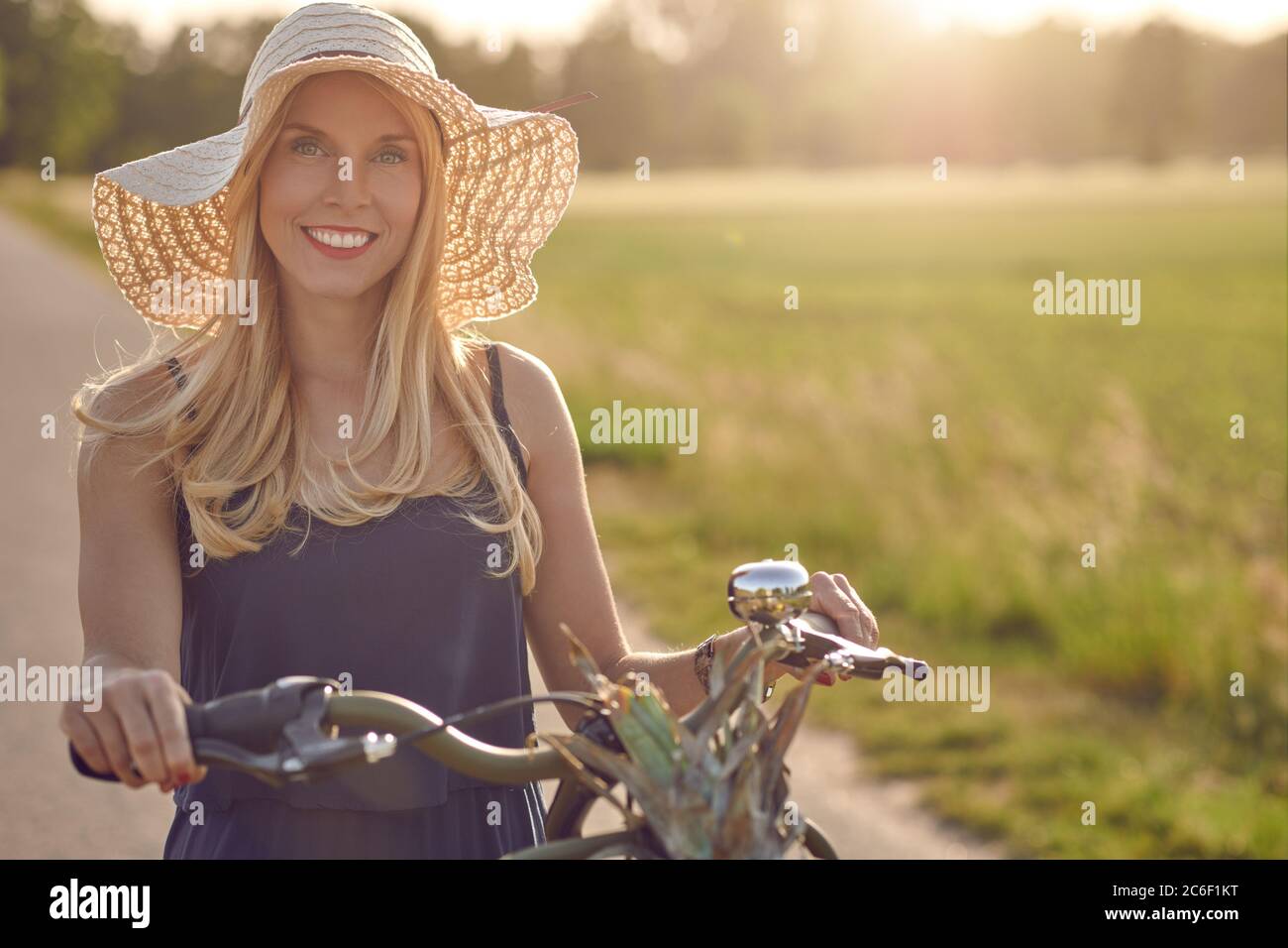 Attraente donna che indossa un ampio cappello di paglia brimmed shopping per prodotti freschi nel paese che ruota la sua bicicletta lungo una corsia rurale in un caldo evenin Foto Stock