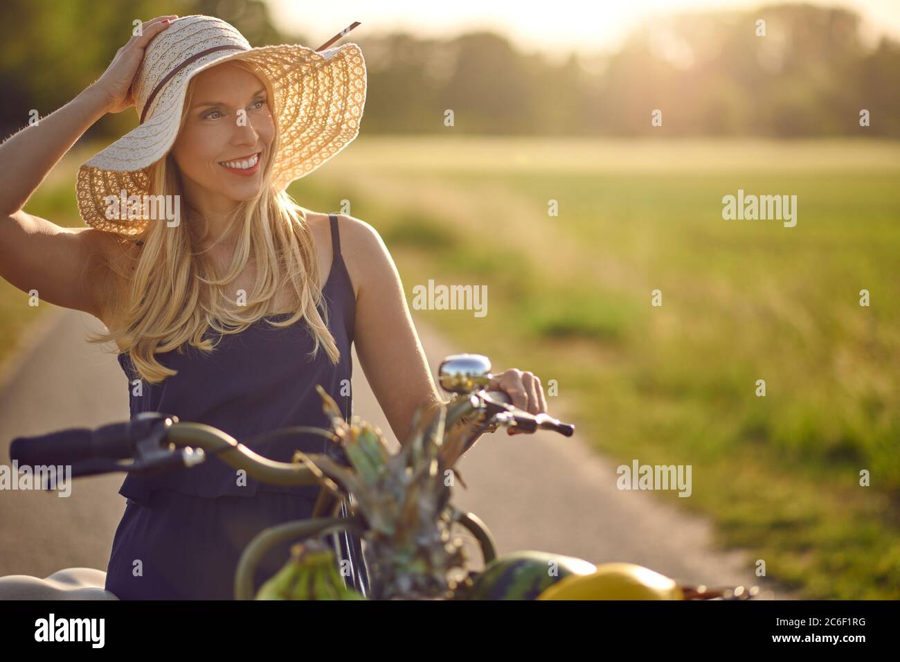 Attraente donna che indossa un ampio cappello di paglia brimmed shopping per prodotti freschi nel paese che ruota la sua bicicletta lungo una corsia rurale in un caldo evenin Foto Stock