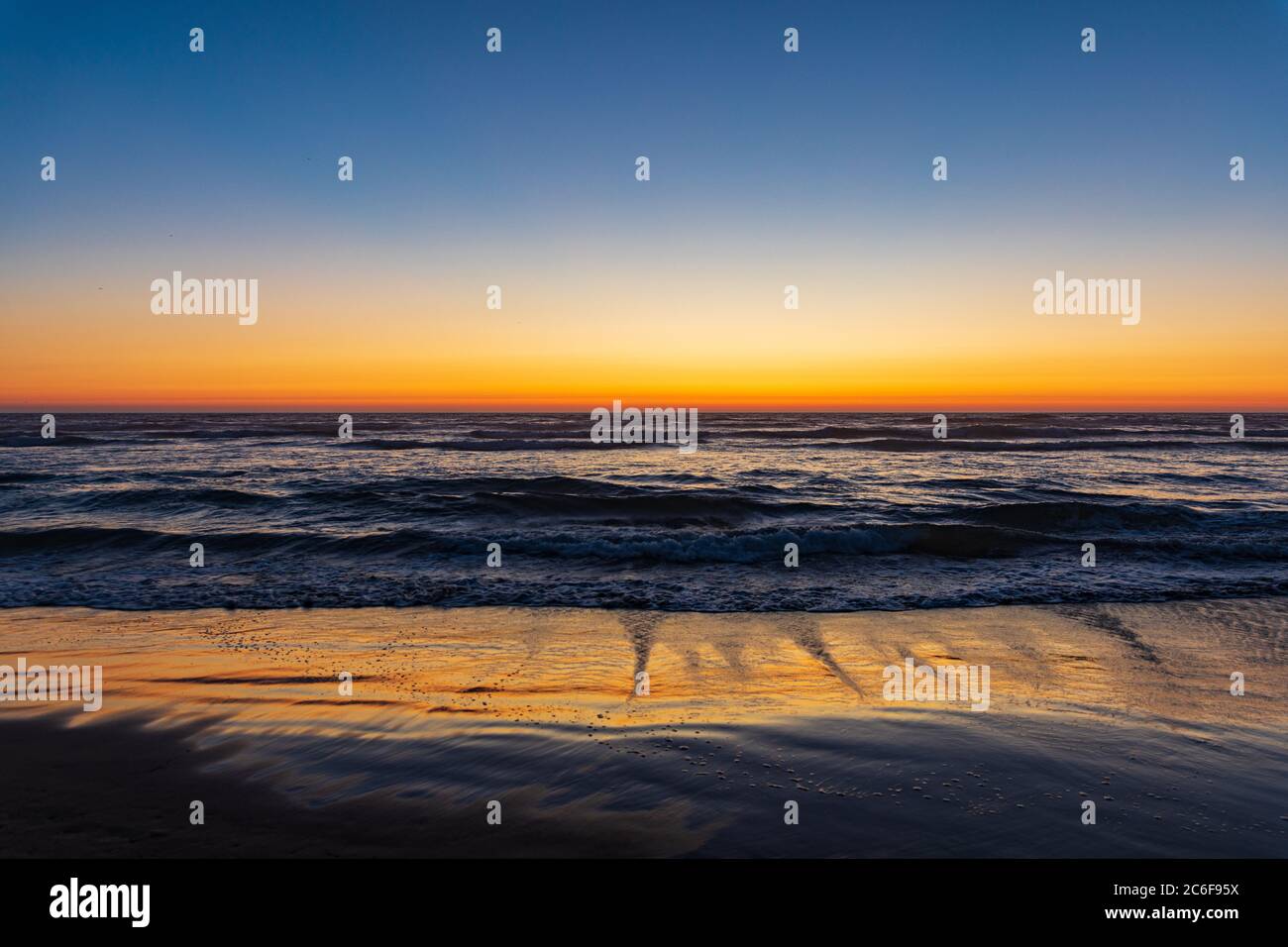 Il tramonto arancione si illumina all'orizzonte e sulla sabbia bagnata mentre le onde si ergono a riva su Cannon Beach, Oregon Foto Stock
