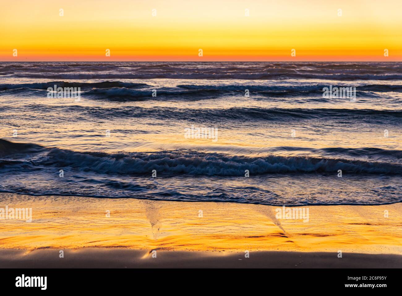 Il tramonto arancione si illumina nel cielo e sulla sabbia bagnata mentre le onde si fermano a riva su Cannon Beach, Oregon Foto Stock