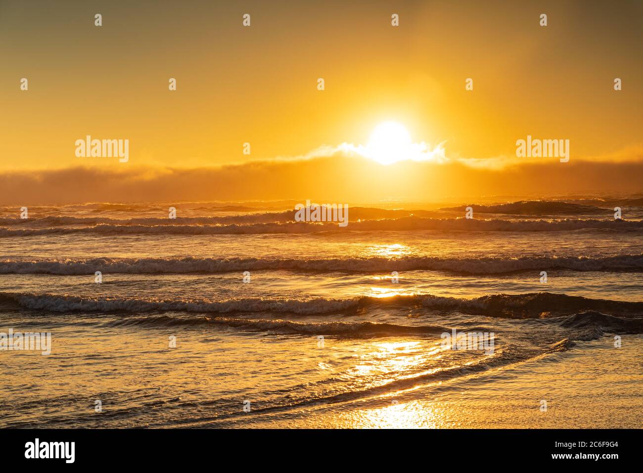 Il grande tramonto arancione trasmette luce dorata attraverso le onde che si infrangono sulla riva. Foto Stock