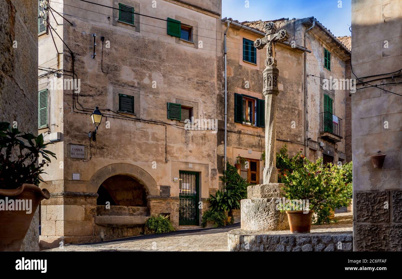 piazza del villaggio mediterraneo chiamata carrer rei sanxo con crocifisso su un palo illuminato dalla luce del sole nel piccolo villaggio di montagna valldemossa Foto Stock