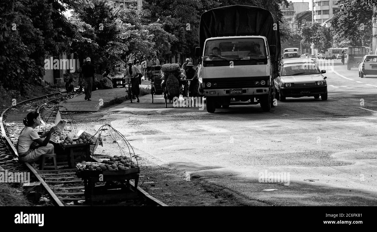 Hawker donna che vende cibo seduto su una pista ferroviaria da una strada trafficata, Yangon, Myanmar, formalmente Birmania, un luogo pericoloso e malsano. Monocromatico Foto Stock