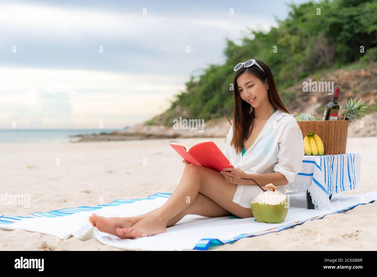 Giovane donna asiatica indossare occhiali da sole è rilassante con la lettura di un libro nella spiaggia di sabbia bianca e vicino al mare con frutta tropicale in background. Estate, holi Foto Stock